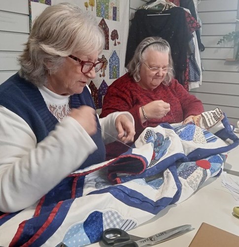 Two Quilts For The Longest Day volunteers sewing a blue, white and red patchwork quilt together.